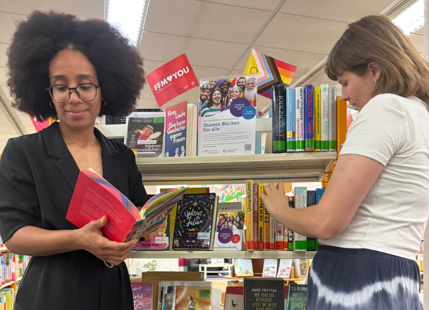 Zwei Personen stehen vor einem Regal mit Büchern. Die Person im Vordergrund hat ein aufgeschlagenes Buch in der Hand.
