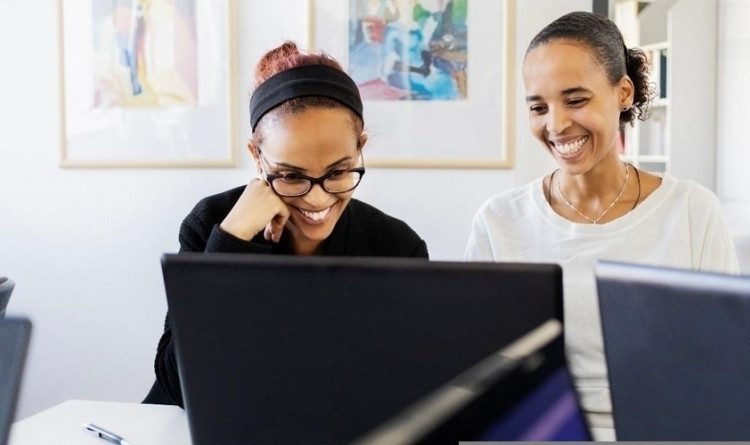 Zwei Frauen sitzen nebeneinander und schauen lächelnd auf einen Laptop. 