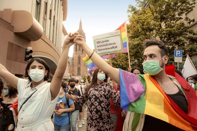 Ausstellungsfoto "Queer refugees at Frankfurt Pride (© Mara Monetti/Stadt Frankfurt) 2 Personen mit Masken demonstrieren auf dem CSD in Frankfurt. Sie haben die Hände gemeinsam in die Höhe gestreckt und tragen eine Regennbogenflagge
