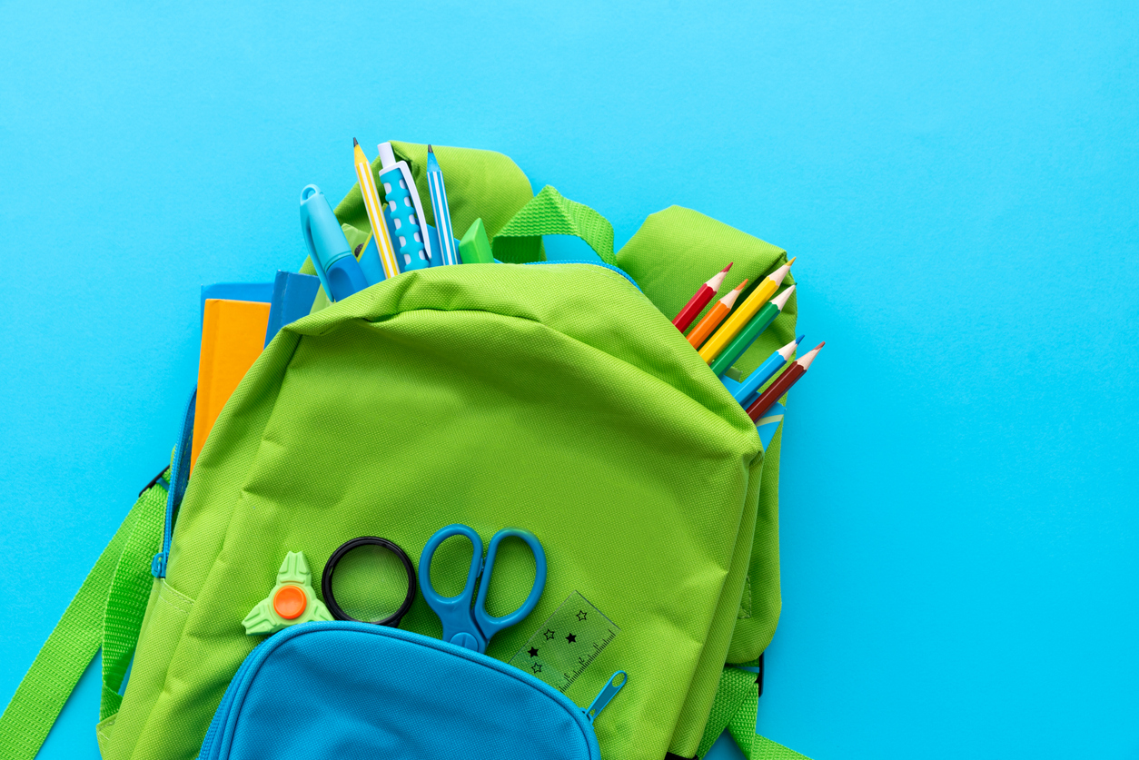 ©istock; Tshum knallgrüner Schulrucksack auf knallblauen Hintergund, aus dem Stifte, Scheren und anderes Schulmaterial rausgucken
