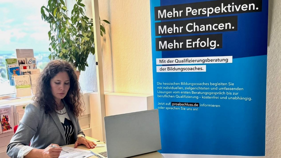 Eine Bildungsberaterin am Schreibtisch neben einem Roll-up zum Programm "ProAbschluss"