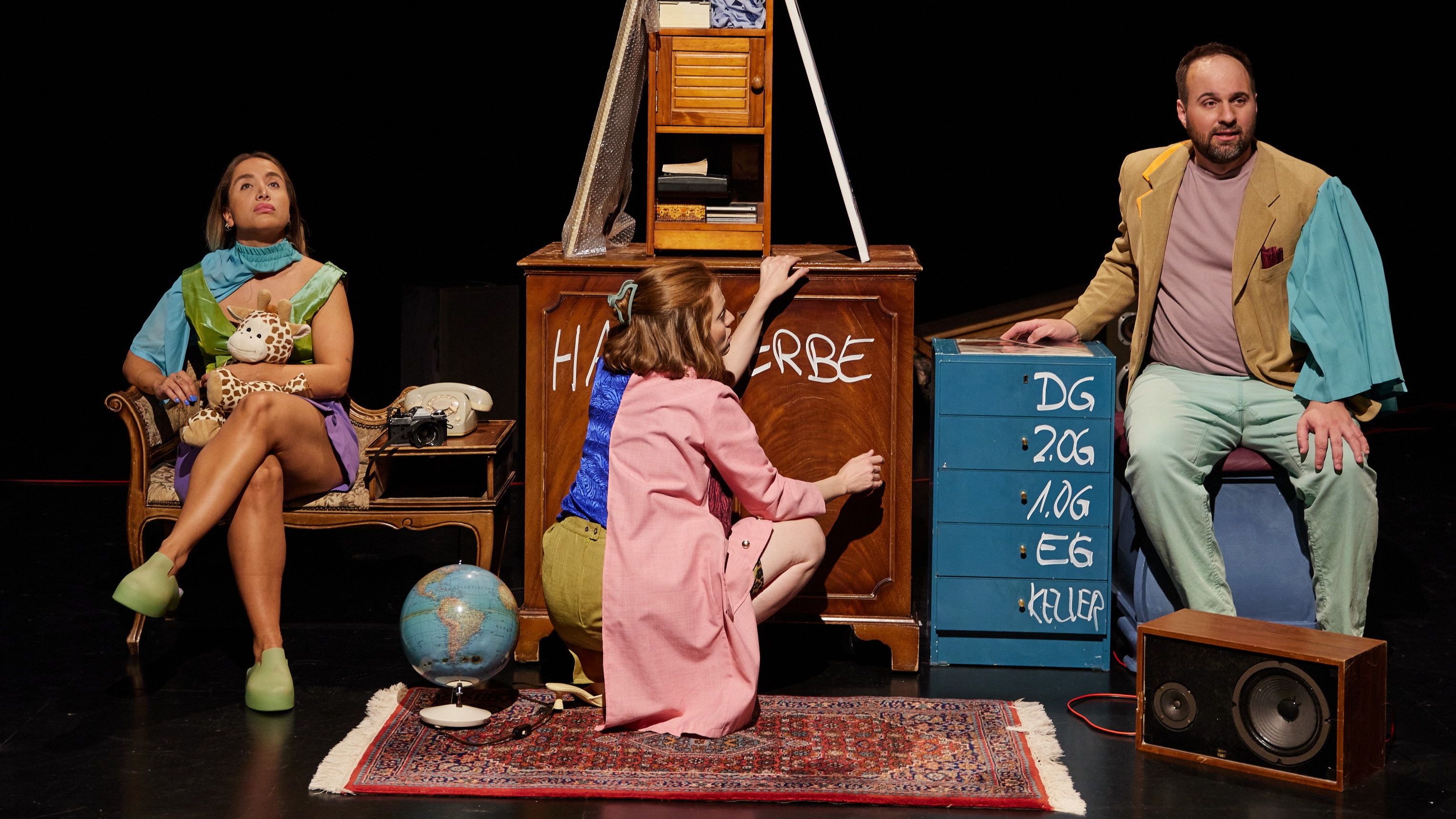 (© Hinterlassenschaft: A House Full of Stuff) Auf einer dunklen Bühne sitzen drei Personen zwischen alten Möbeln und Alltagsgegenständen. Links hält eine Frau mit buntem Outfit und grünen Schuhen ein Stofftier im Arm und blickt nach oben. In der Mitte kniet eine Frau im rosa Mantel vor einem Schrank mit der Aufschrift „HINTERERBE“. Rechts sitzt ein Mann in heller Kleidung neben einem blauen Schubladenschrank, der mit verschiedenen Stockwerken beschriftet ist. Vor ihnen liegen ein Teppich, ein Globus, eine Brille und eine Lautsprecherbox.
