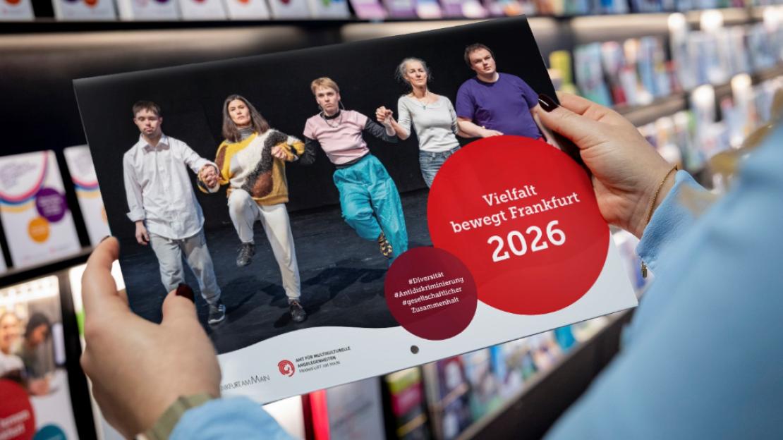 Der "Vielfalt bewegt Frankfurt"-Kalender 2026 in den Händen einer Person vor einem Flyerregal