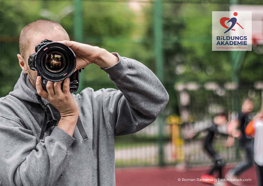 Zu sehen ist ein Mensch in einem grauen Pullover, der eine Kamera auf die betrachtende Person richtet. Im Hintergrund verschwommen Sportler:innen auf einem Sportplatz. Logo Bildungsakademie des lsb h e.V.
