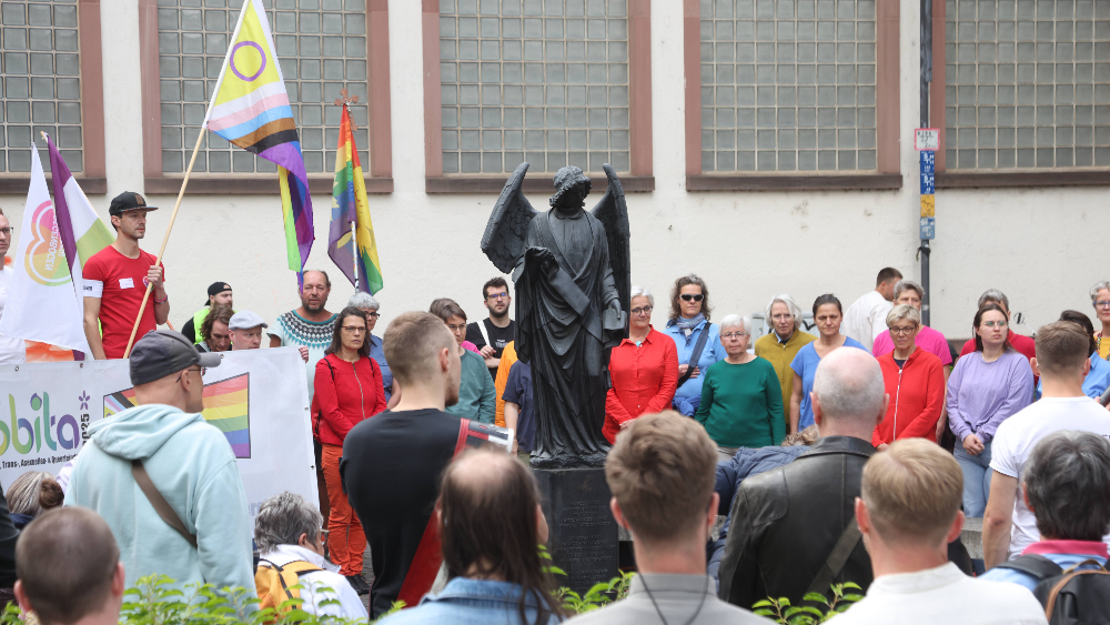 Eine Gruppe von Personen steht beim IDAHOBITA* 2025 mit Regenbogenfahnen zum Gedenken um das Mahnmal "Frankfurter Engel". 
