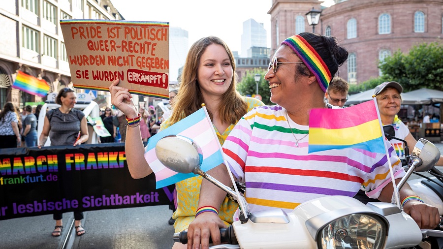 Zwei weiblich gelesene Personen sitzen auf einem Roller und lächeln sich an.