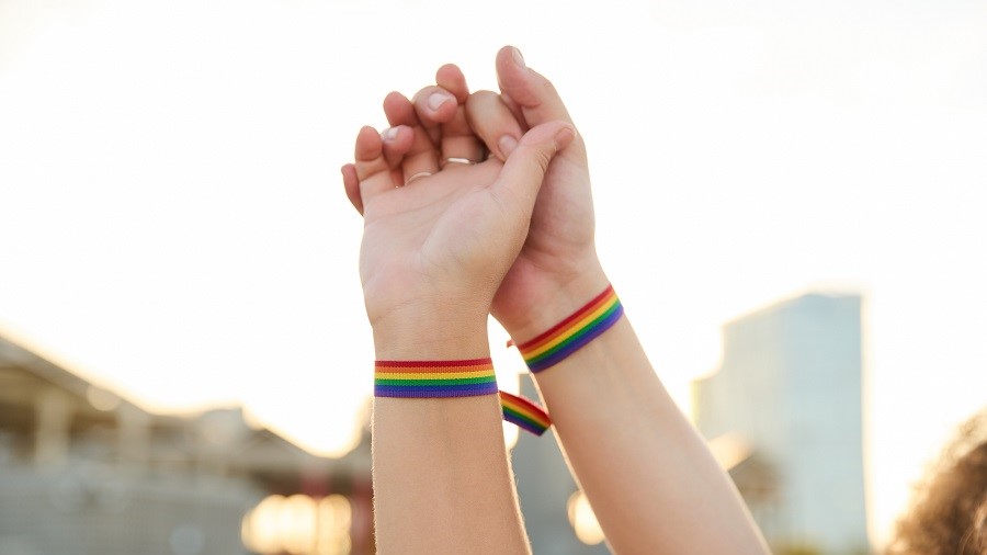 Zwei Hände berühren sich. An den Handgelenken ist ein Pride-Bändchen.
