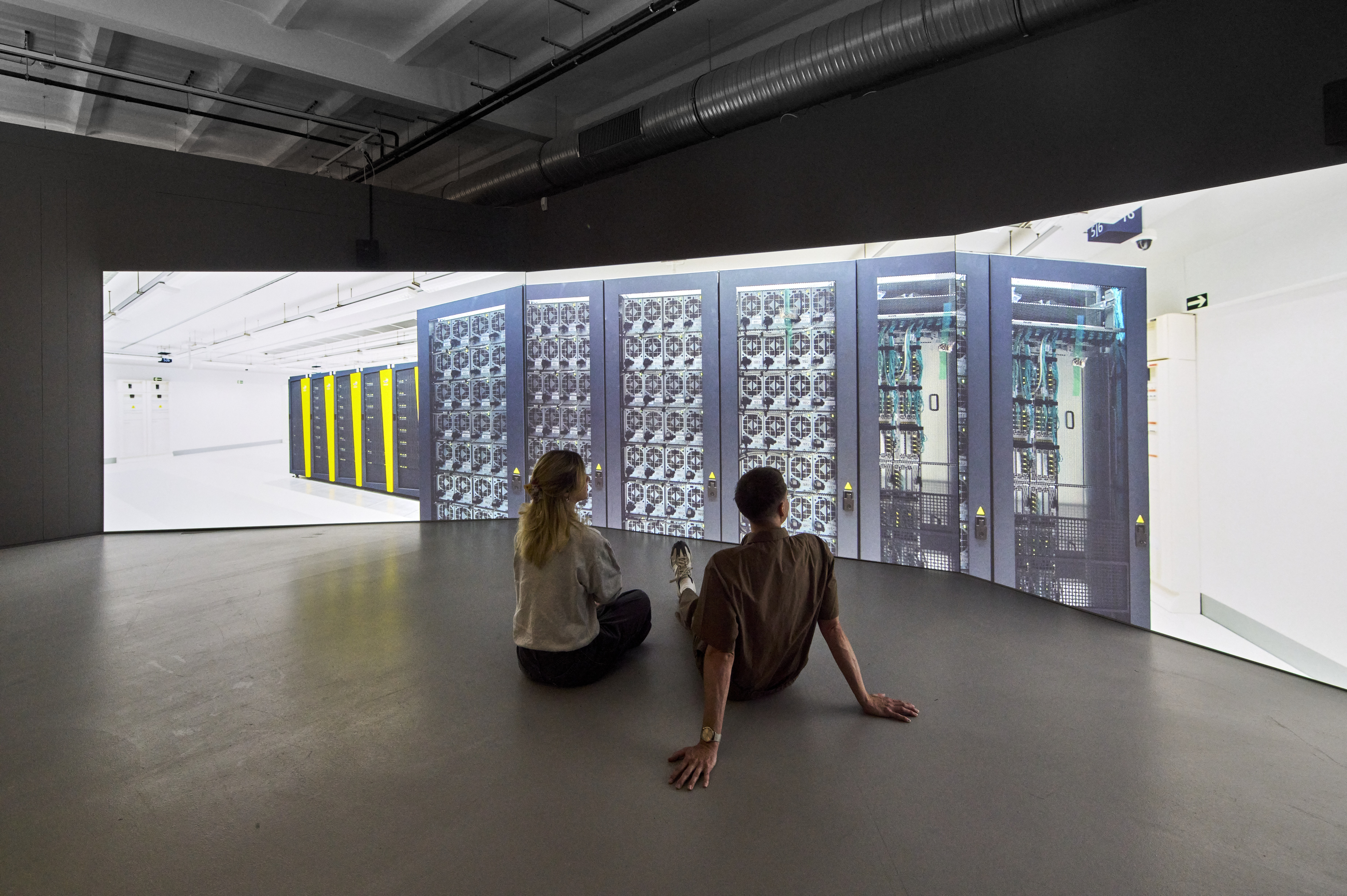 Zu sehen sind eine weiblich und eine männlich gelesene Person, die auf dem Boden des Ausstellungsraumes sitzen und eine Videoarbeit anschauen. Auf den Bildschirmen ist ein komplett weißer Raum zu erkennen, in dem KI-Datenserver aufbewahrt werden.