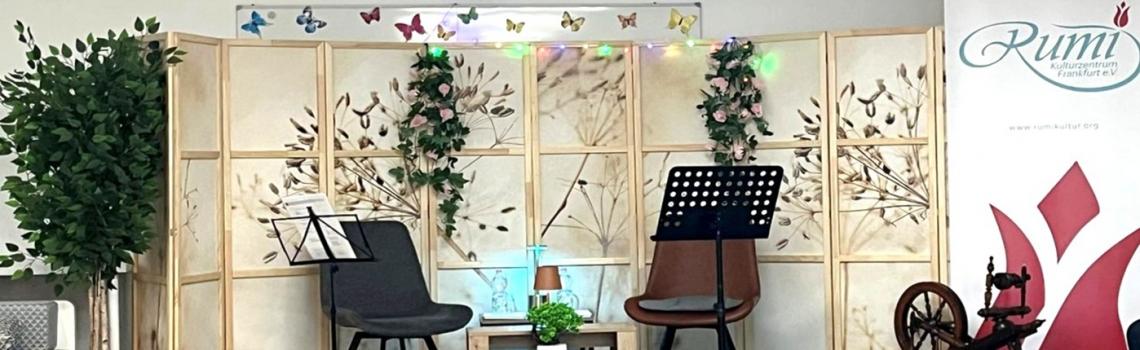 Eine Bühne mit zwei Stühlen, weiteren Gegenständen und einem Roll-up mit dem Rumi-Logo (© Rumi Kulturzentrum Frankfurt)